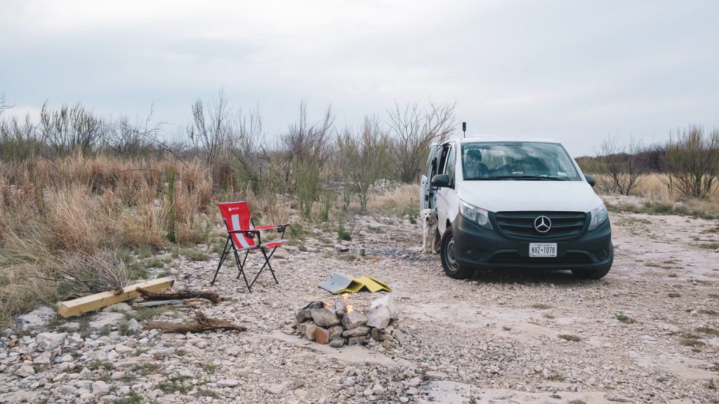 Van and campfire with red chair, in a clearing on rocks with desert brush around