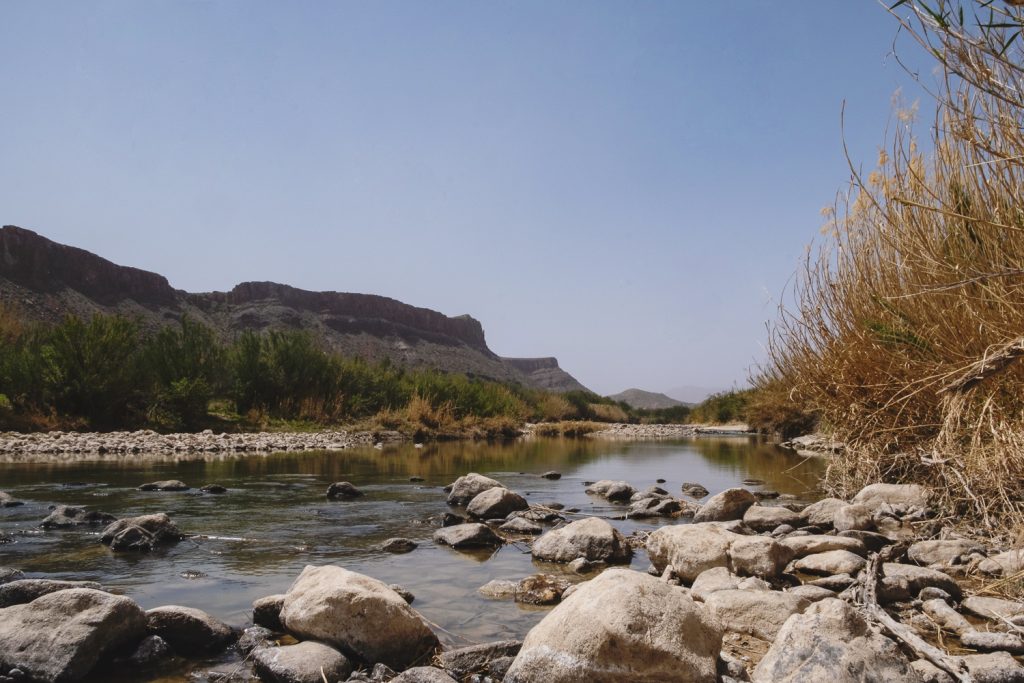 View of the gentle Rio Grande River with rocks in the water, grasses on the right and green shrubs and mountains rising on the left