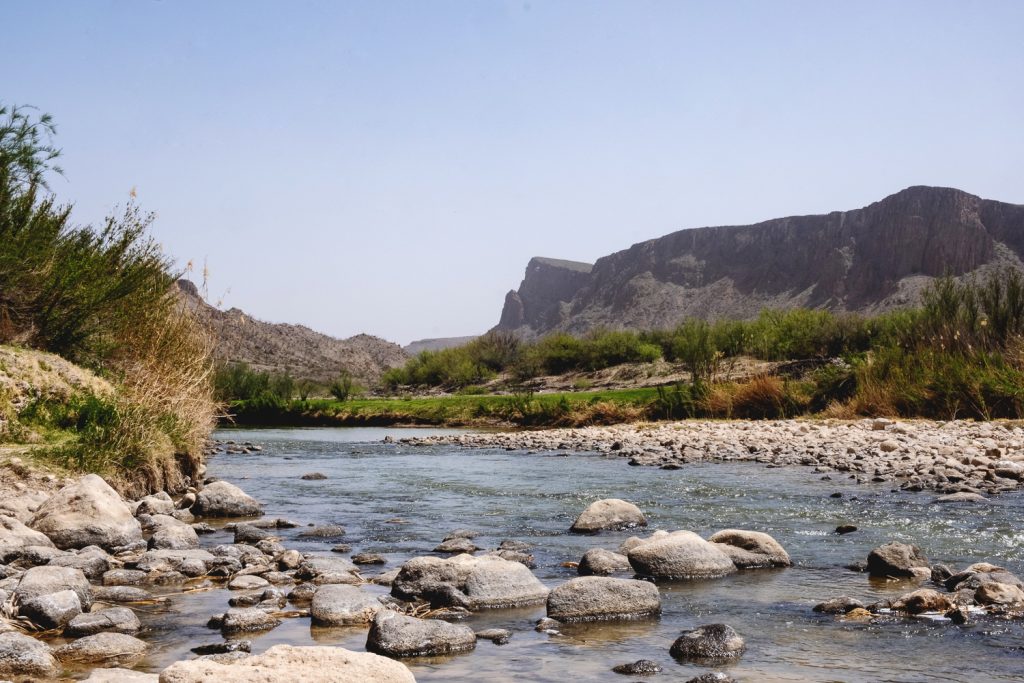 View of the gentle Rio Grande River with rocks in the water, grasses, green shrubs and mountains rising 