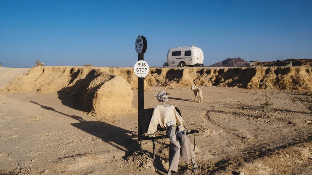 Random fake bus stop on a desert road with a skeleton on the bench and mesa and camper behind