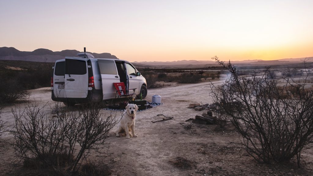 Campsite with the white van, red chair, Zeke and smoking campfire, on the white desert mesa with the sun setting in the distance mountains.