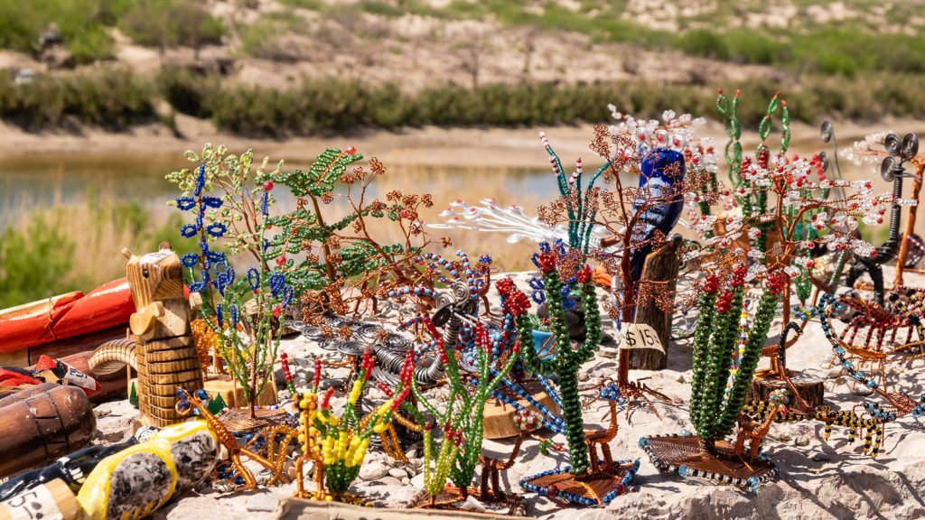 Colorful trinkets made from beds sit atop a rock, with the Rio Grande and Mexican border behind