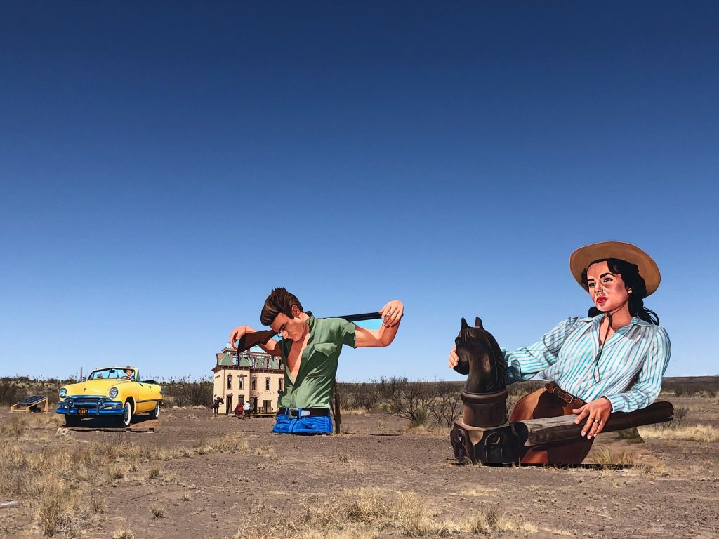 Random road-side  attraction with larger than life cutout billboards... car, house, dude with a gun, and cowgirl on a horse that's a chest piece... blue sky